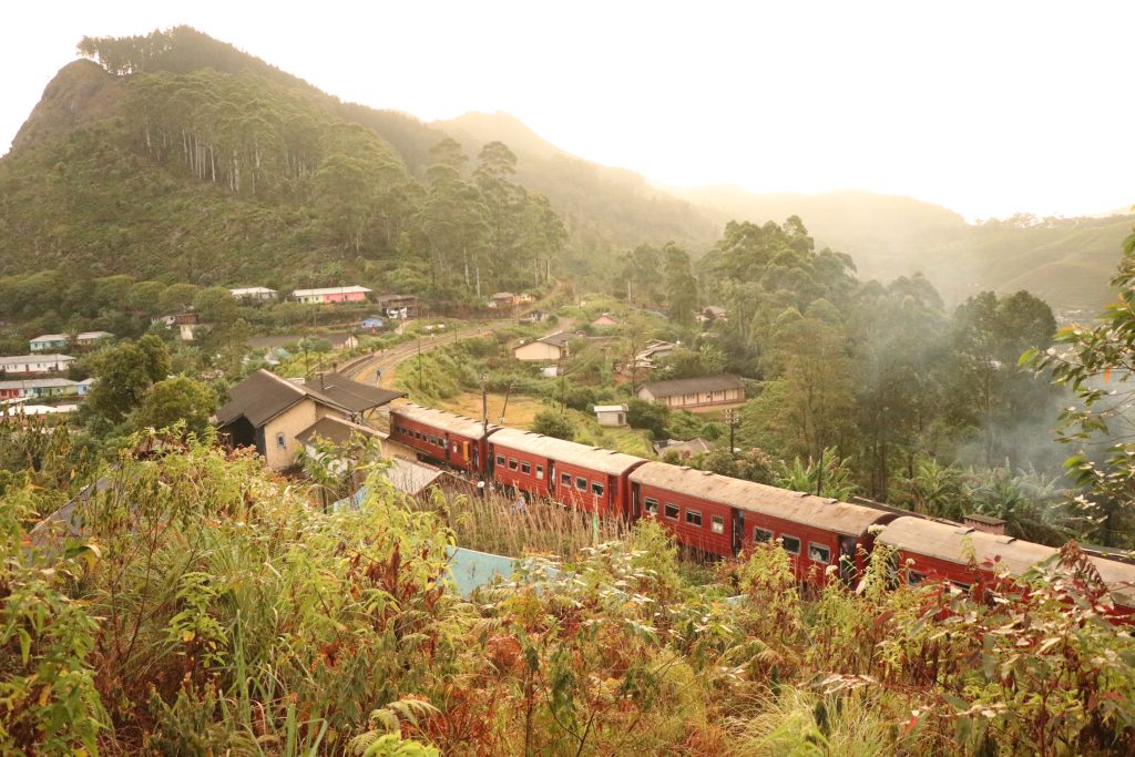 A red train winding along the hillside below a village at Idalgashinna, with forested hills and morning haze behind