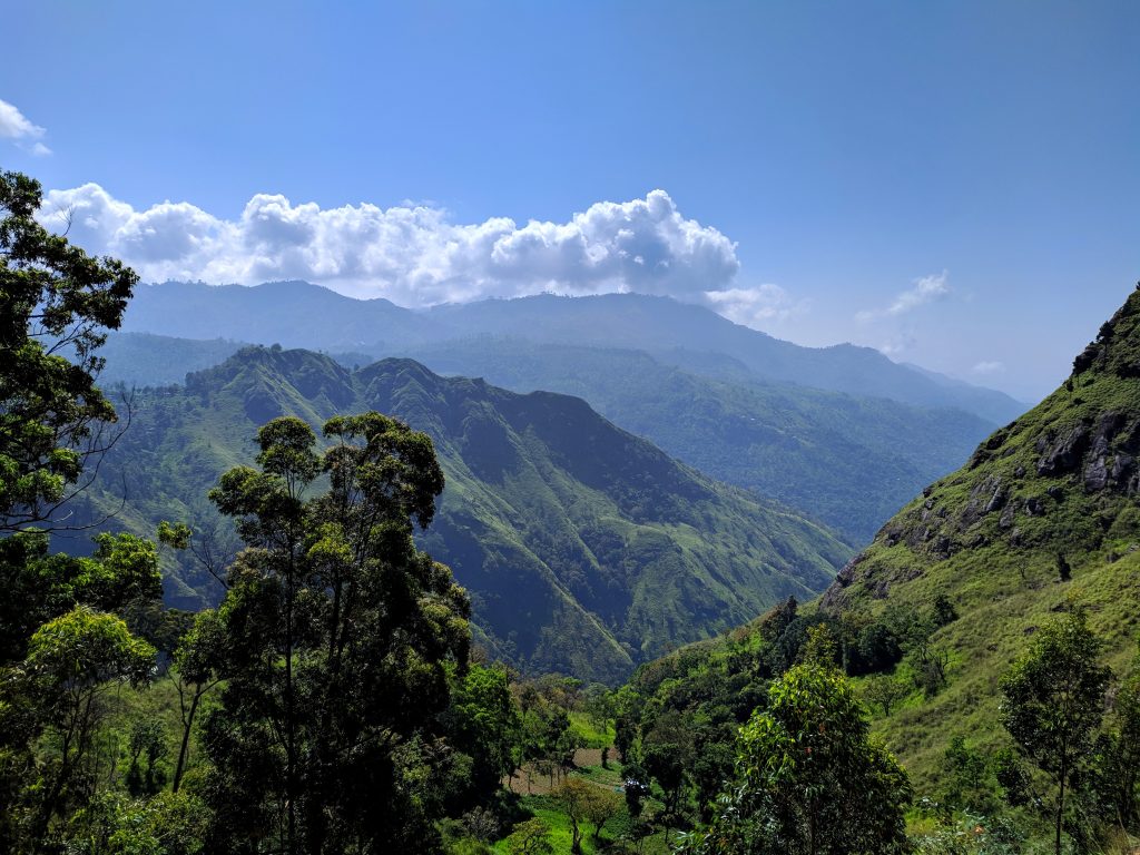 Steep forested hills and a deep green valley under a blue sky with white clouds, in the hill country around Ella
