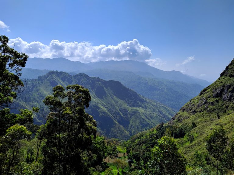 Steep forested hills and a deep green valley under a blue sky with white clouds, in the hill country around Ella