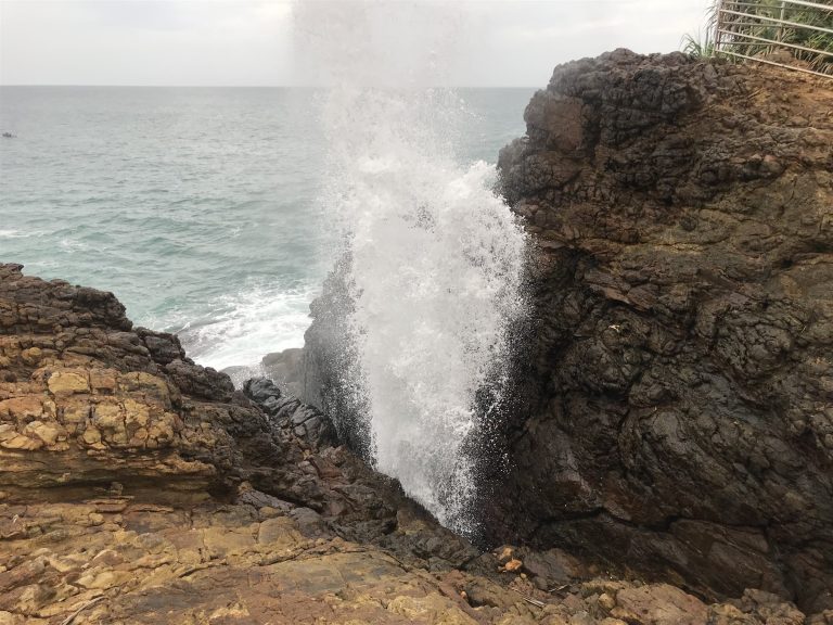 A tall column of white spray rising from a rocky cliff-top gap at Hummanaya blowhole, with the sea behind and a safety railing at top right