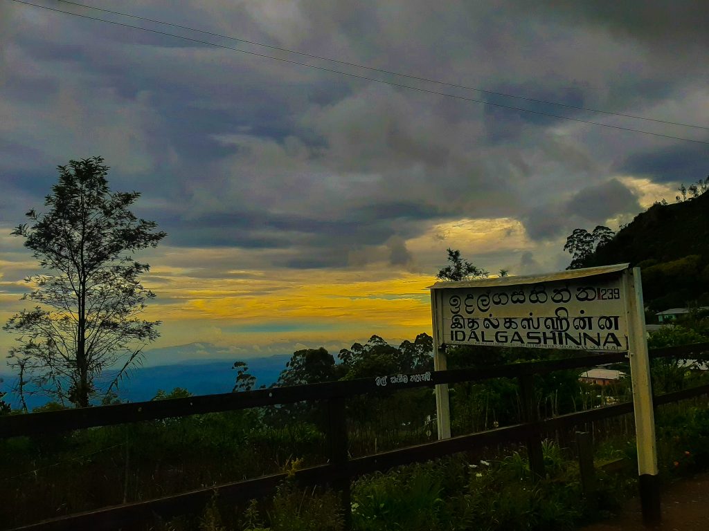 The Idalgashinna station name board in Sinhala, Tamil and English beside a wooden fence, with hills falling away to distant lowlands under a cloudy evening sky