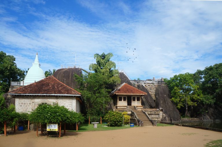 Isurumuniya temple: a tiled shrine with steps set against dark granite boulders, a white stupa on the left and a pond at the right edge