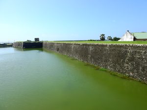 The long stone rampart of Jaffna Fort rising from a green moat under a clear blue sky, with a colonial building on the grassy wall top
