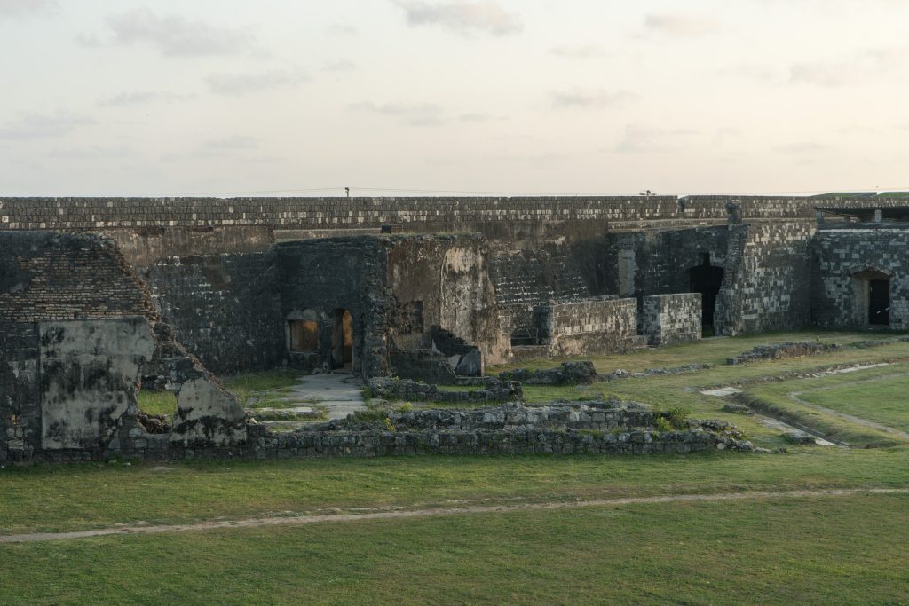 Roofless coral-stone ruins inside Jaffna Fort below the long rampart wall, with a lawn in the foreground