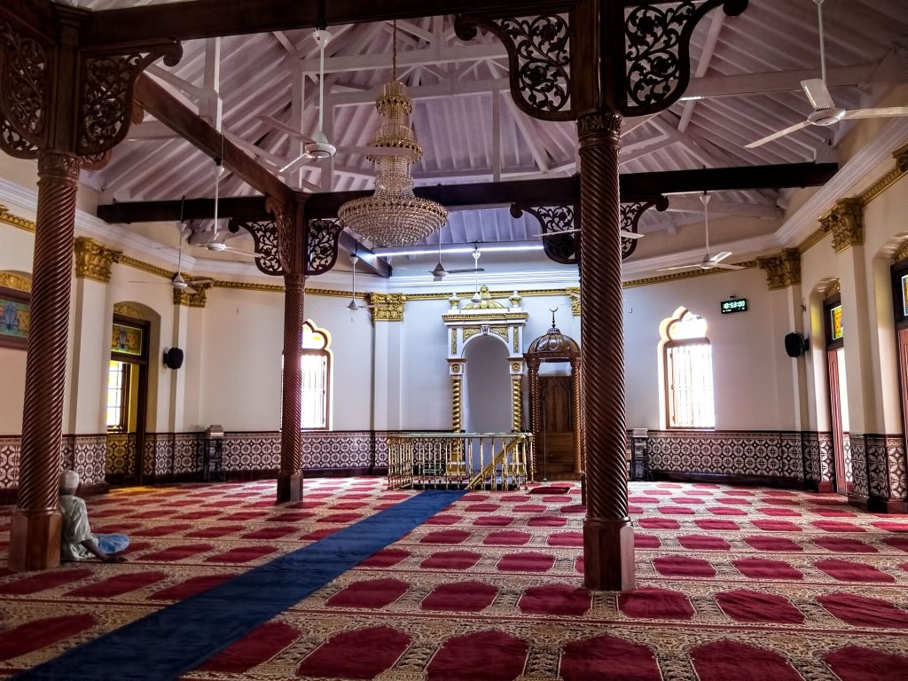 The upstairs prayer hall of the Jami Ul-Alfar Mosque with carved wooden pillars, a crystal chandelier and red prayer carpets