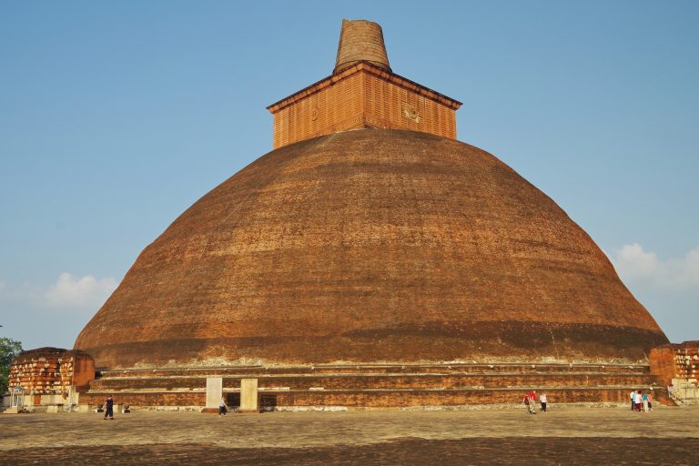 The huge bare-brick dome of Jetavanaramaya with a square top and short spire, above a wide paved platform with a few visitors