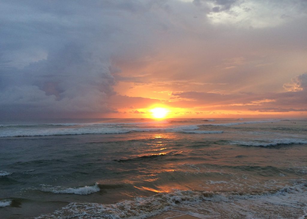 Sunset over the sea at Kabalana Beach, with waves breaking on the reef and surfers waiting in the water on the right