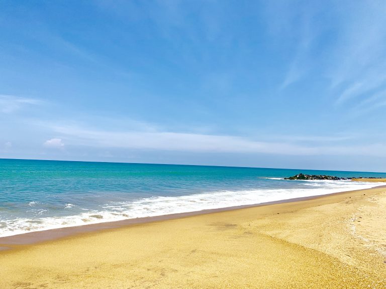 Wide golden beach at Kalutara with small waves and a rock groyne in the distance under a blue sky