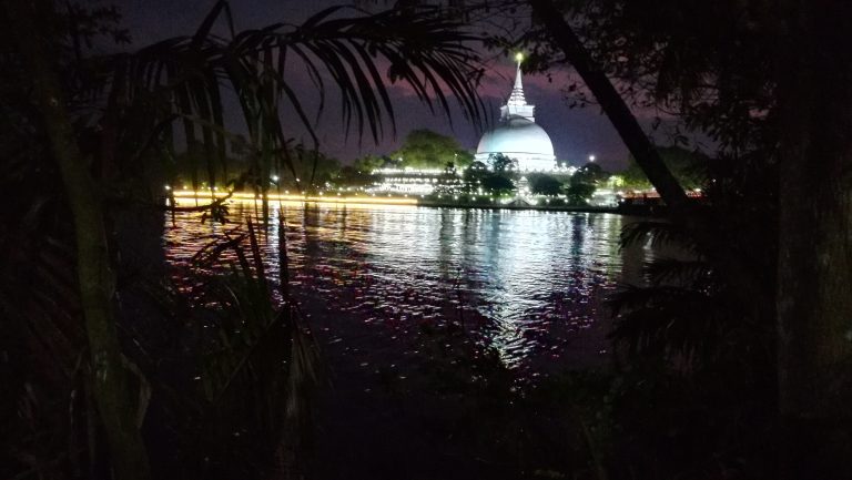 The Kalutara stupa lit up at night, its lights reflected in the Kalu Ganga, seen through palm fronds from the far bank