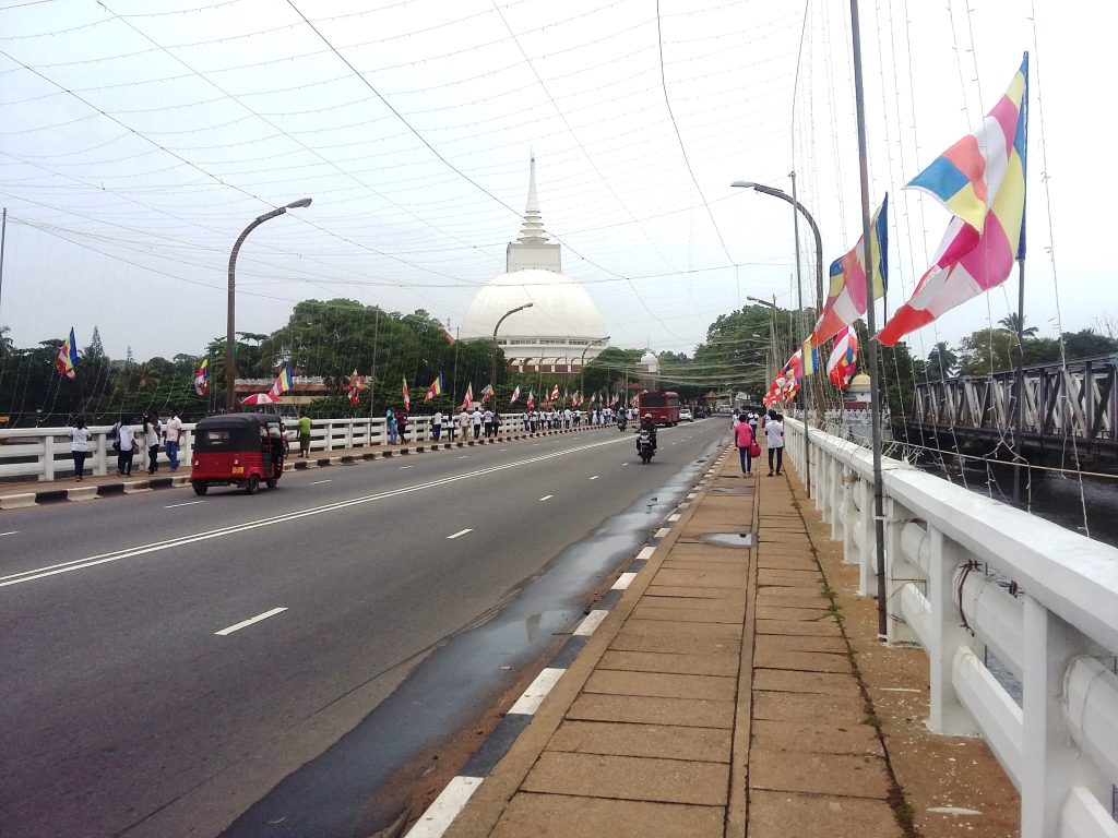 Galle Road crossing the Kalutara bridge towards the white stupa of the Kalutara Bodhiya, with Buddhist flags along the railings and a tuk-tuk on the road
