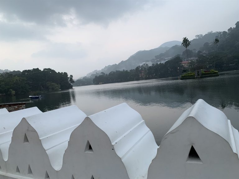 White scalloped wall with triangular openings beside Kandy Lake on a misty morning, wooded hills beyond
