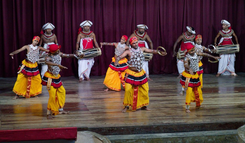 Dancers in red, white and yellow costumes and headdresses performing with drums on a stage in Kandy, in front of a red curtain