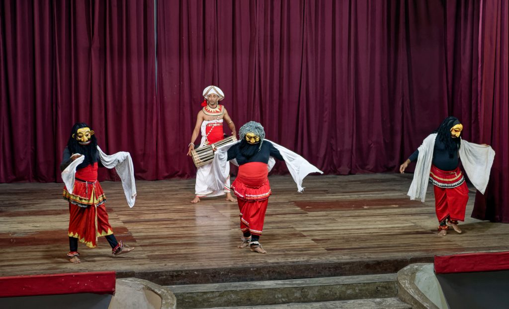 Masked dancers in red and white costumes waving white cloths on stage, with a drummer in a white headdress behind