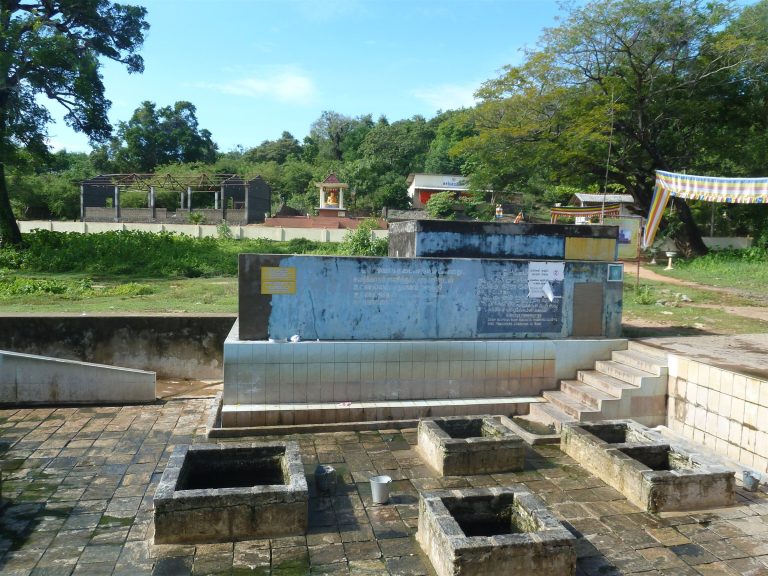 Four stone-framed hot wells in a tiled courtyard at Kanniya with a tiled wall and trees behind