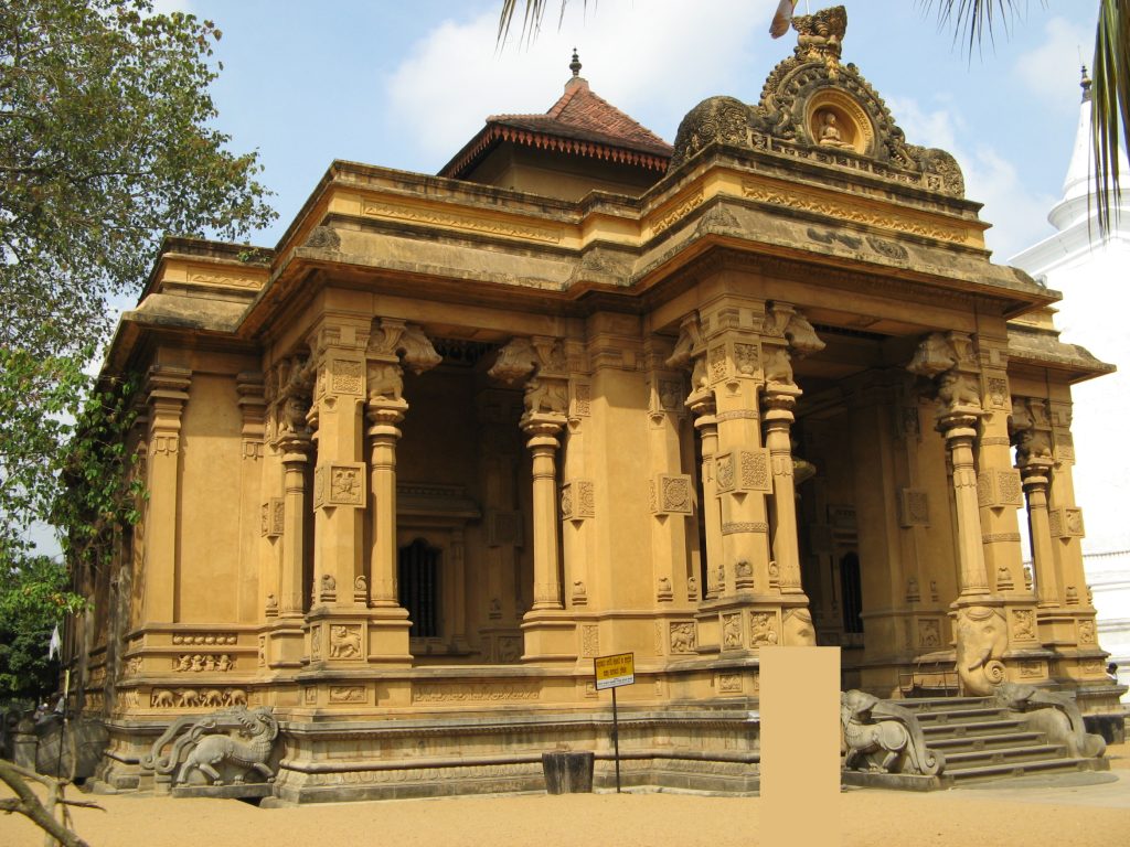 The ochre front of the Kelaniya image house, with carved columns, an ornamented gable and guardian lions beside the steps
