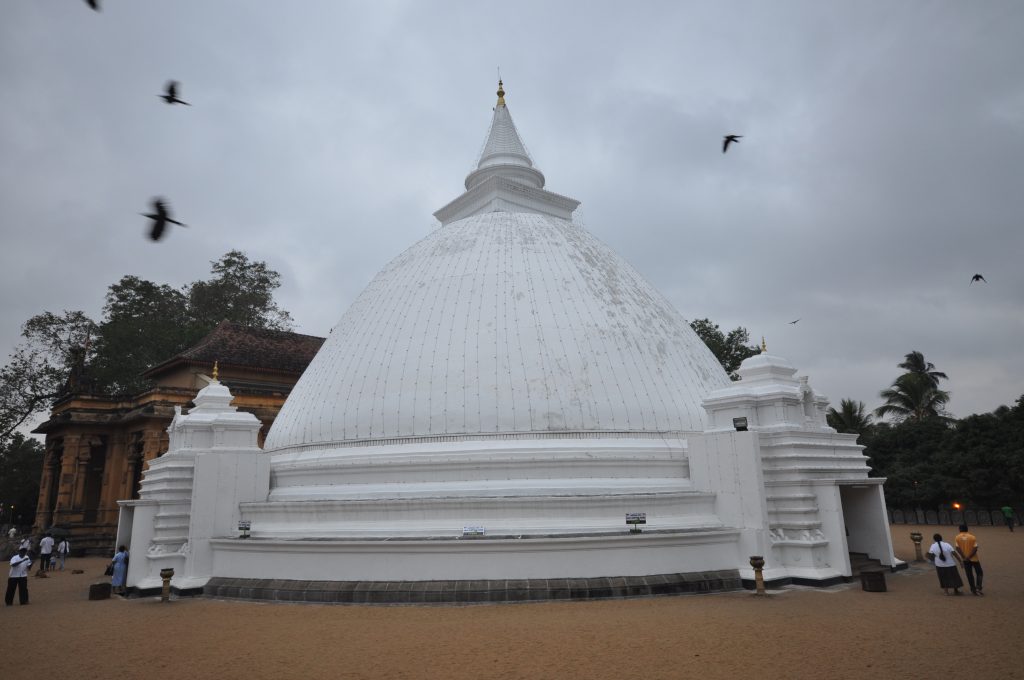 The large white paddy-heap stupa of Kelaniya temple with small gateway shrines at its base, rising above a sandy courtyard under a cloudy sky