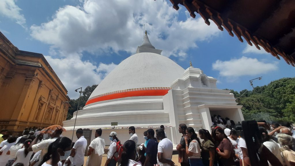Crowds of worshippers, many in white, walking around the white stupa at Kelaniya Raja Maha Vihara under a blue sky