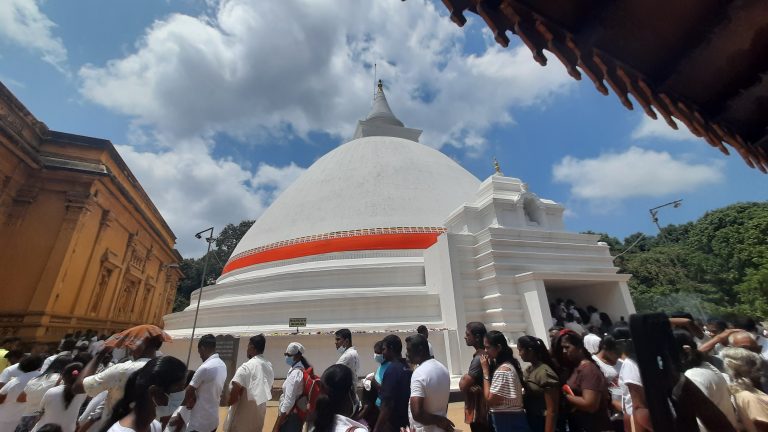 Crowds of worshippers, many in white, walking around the white stupa at Kelaniya Raja Maha Vihara under a blue sky