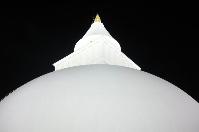 Looking up at the white dome, square harmika with a carved wheel and the gilded finial of Kiri Vehera against the night sky