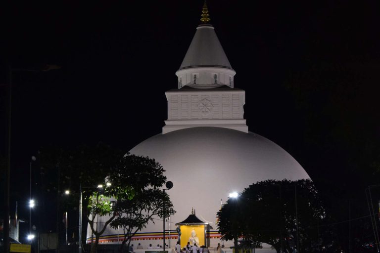 The floodlit white dome and spire of Kiri Vehera at night, with devotees at the Buddha niche at its foot