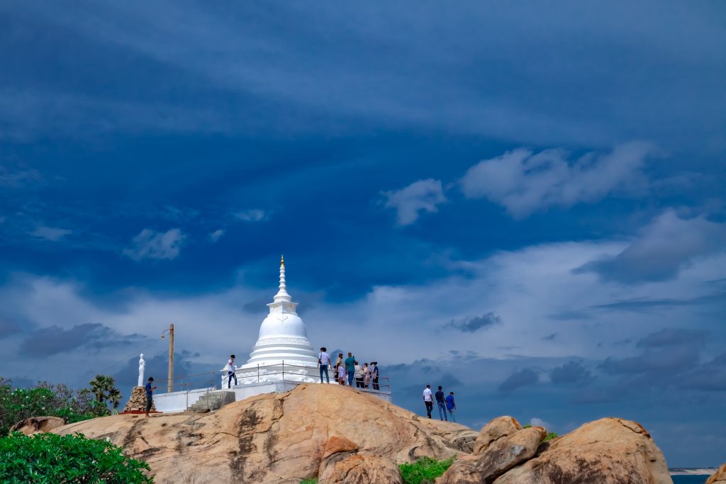 The white dagoba of Kirinda Temple on its rocky outcrop above the sea, with a white statue of Vihara Maha Devi to one side and a dramatic sky behind