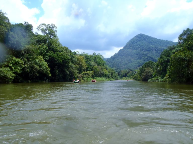 A wide, calm stretch of the brown Kelani River between forested banks, with a forested hill ahead and rafts small in the distance
