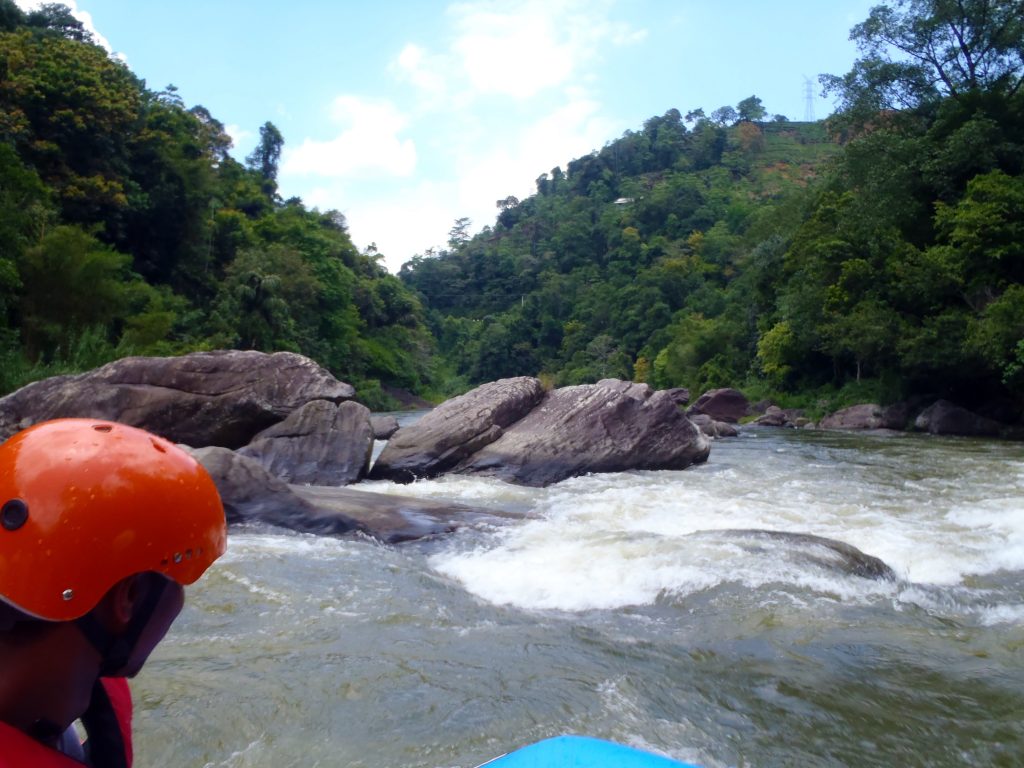 View from a raft on the Kelani River at Kitulgala: white water breaking over grey boulders ahead, forested hills on both banks and a guide's orange helmet in the foreground
