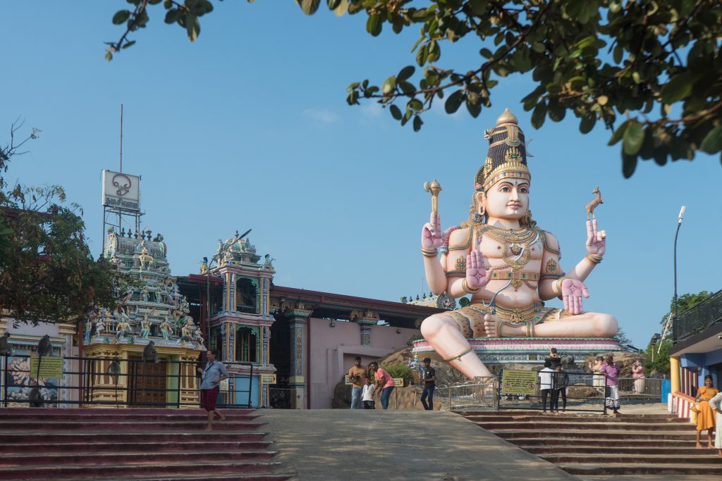 Koneswaram Temple's carved gateway tower and a giant seated Shiva statue at the top of a wide flight of steps, under a clear blue sky