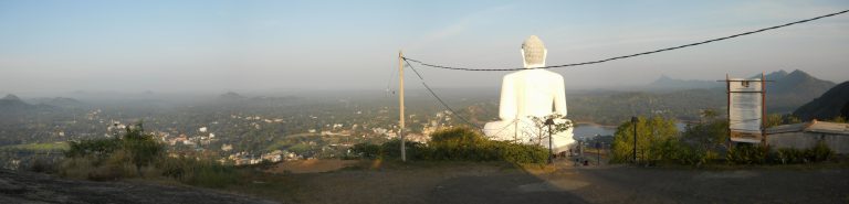 Panorama from the top of Ethagala: the white Buddha statue seen from behind, with Kurunegala town and distant rocks spread out below in the morning haze