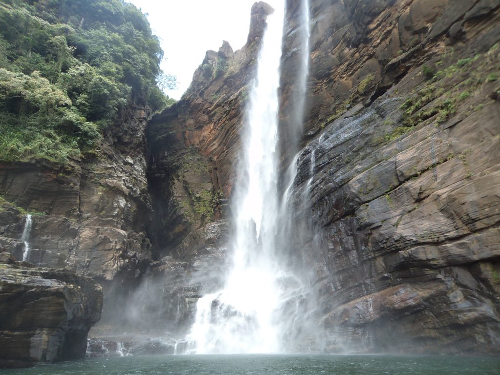 Laxapana Falls in cloud, a thin white stream falling down a cliff above rocks and thick green vegetation