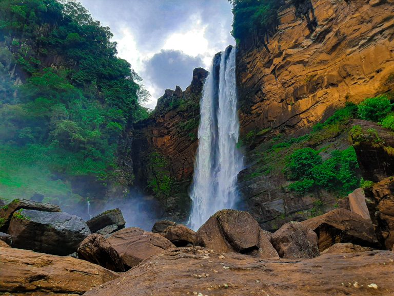 Laxapana Falls dropping down a tall orange-brown rock face beside green forest, seen from the boulders at its base