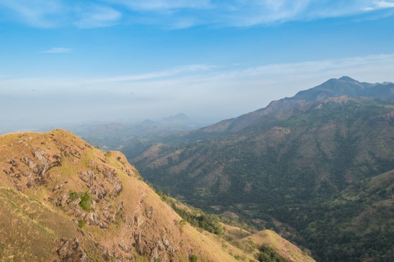 View from the top of Little Adam's Peak over a rocky grass slope and a deep green valley towards hazy lowlands