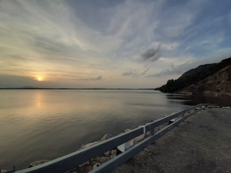 Calm reservoir at sunset beside a roadside railing, with a forested rocky hill on the right