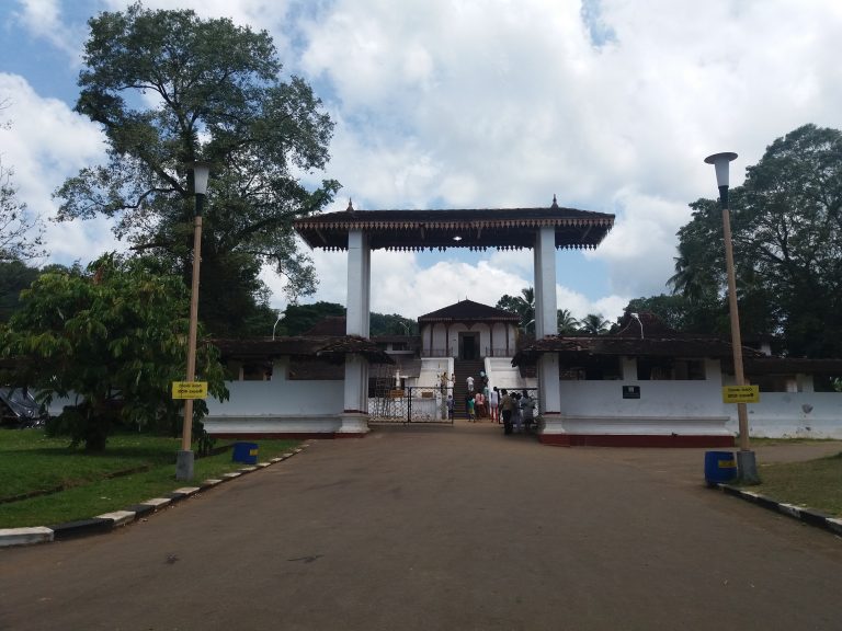 The white pillared entrance gateway of the Maha Saman Devalaya in Ratnapura, with steps leading up to the shrine and tall trees on either side