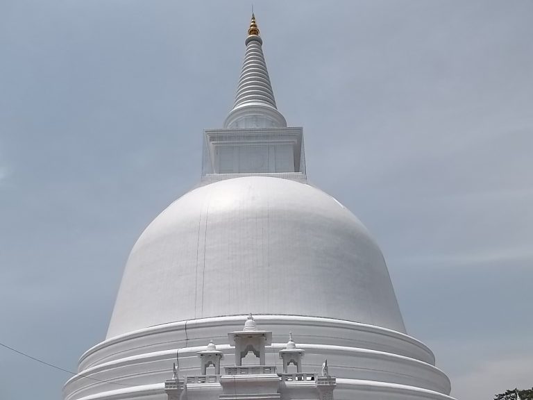 The white dome and tall spire of the Mahiyangana stupa against a grey sky, with small white shrines on its base