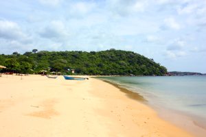 A curving sandy beach with two fishing boats and thatched sunshades, calm turquoise water and a forested headland ending in rocks