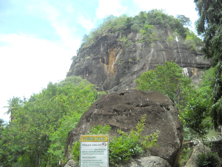 The dark, streaked rock face of Mulkirigala rising above trees, with a large boulder and a temple notice board in front