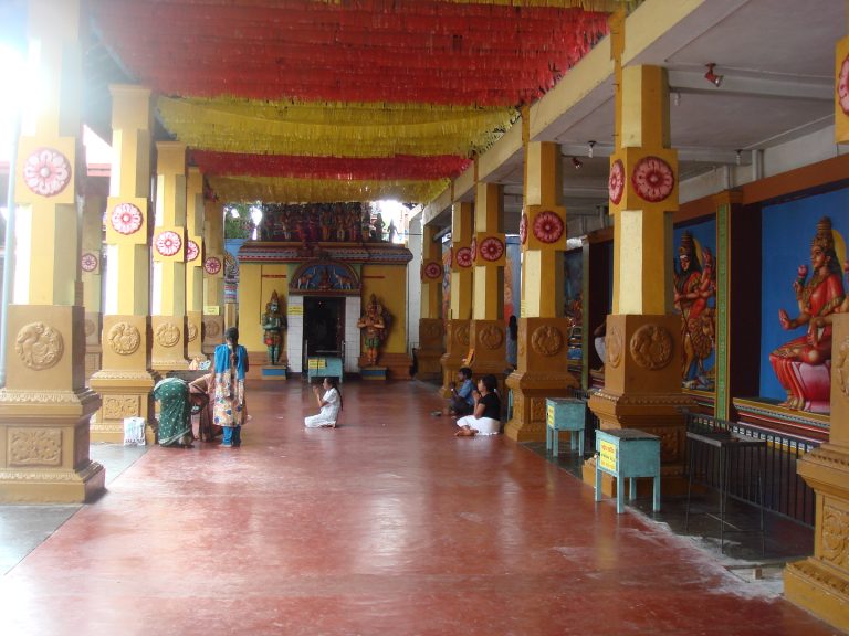 Pillared hall at Munneswaram temple with red and yellow streamers overhead, painted deities on the walls and worshippers praying in front of a small shrine