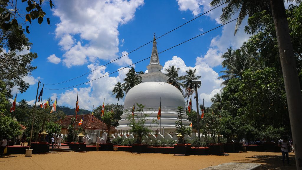 The white stupa of Muthiyangana Raja Maha Vihara in Badulla under a blue sky, with Buddhist flags, palm trees and a sandy courtyard in front