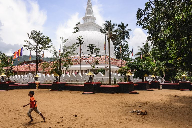 The white Muthiyangana stupa behind a low crenellated wall hung with Buddhist flags, seen across a sandy courtyard with trees and wooded hills behind