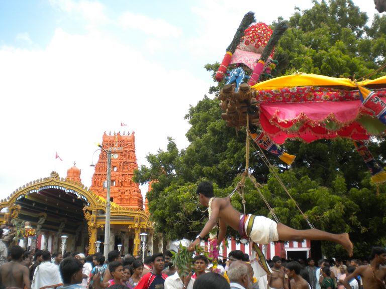 Festival crowd outside Nallur Kandaswamy Temple, with the orange gopuram and golden entrance arch behind; a devotee hangs horizontally from hooks under a decorated kavadi frame above the crowd