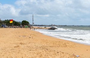 Negombo beach with golden sand, a red-and-yellow flag, waves breaking over a pile of rocks and the town's hotels in the distance under a cloudy sky