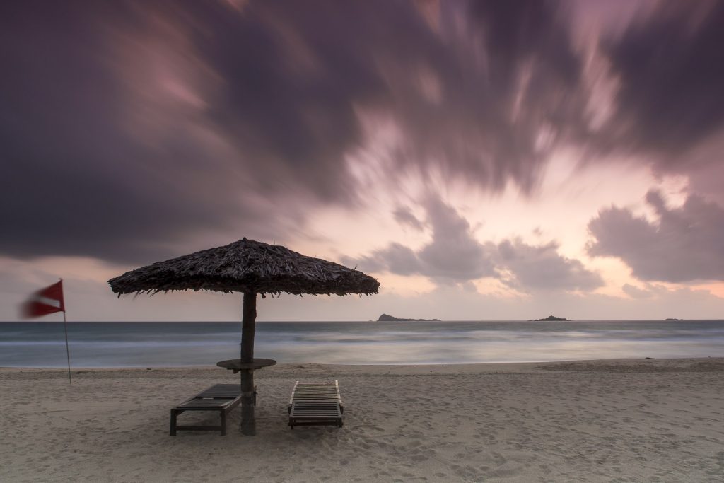 A palm-leaf sunshade, two sun loungers and a red flag on the sand at Nilaveli at dawn, with long-exposure clouds streaking over the sea and small islands on the horizon