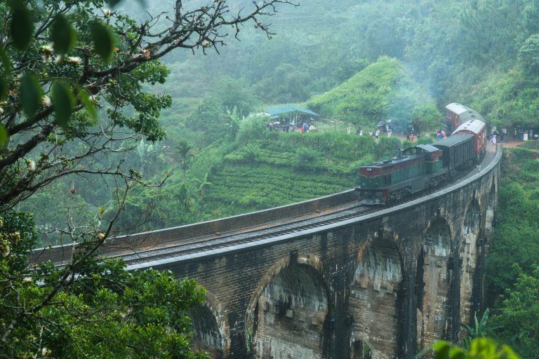 A diesel train with red carriages crossing the stone arches of Nine Arch Bridge near Ella, with tea fields and onlookers on the hillside behind