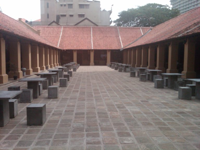 A paved courtyard at the Old Dutch Hospital in Colombo, with low tile-roofed verandas on either side and stone tables and stools
