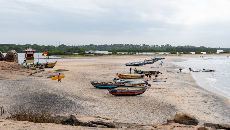 Wide sandy beach with fishing boats pulled up on the sand, surfers carrying boards to the water, a lifeguard tower with a red and yellow flag on the left and a lagoon behind