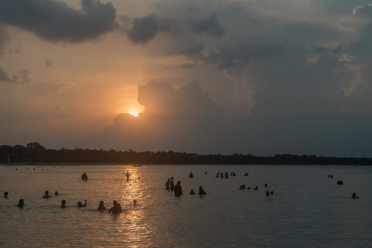 Bathers silhouetted far out in the calm, shallow water of Pasikuda bay at sunset, with a tree on the shore