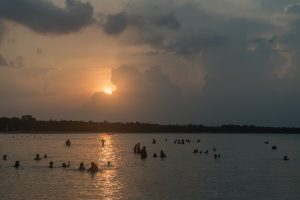 Bathers silhouetted far out in the calm, shallow water of Pasikuda bay at sunset, with a tree-lined shore on the horizon