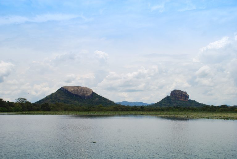 Two forested rocks across a lake: the long flat-topped Pidurangala on the left and the sheer-sided Sigiriya rock on the right