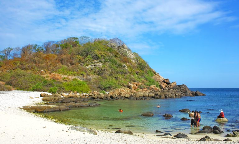 A white beach and turquoise cove on Pigeon Island below a scrub-covered rocky hill, with snorkellers in the shallow water